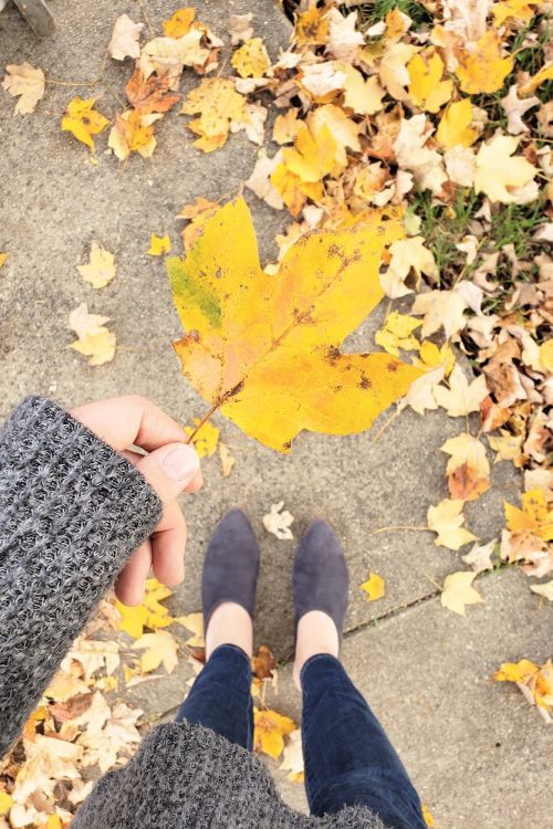Woman holding a yellow leaf towards ground with other leaves spread around sidewalk.
